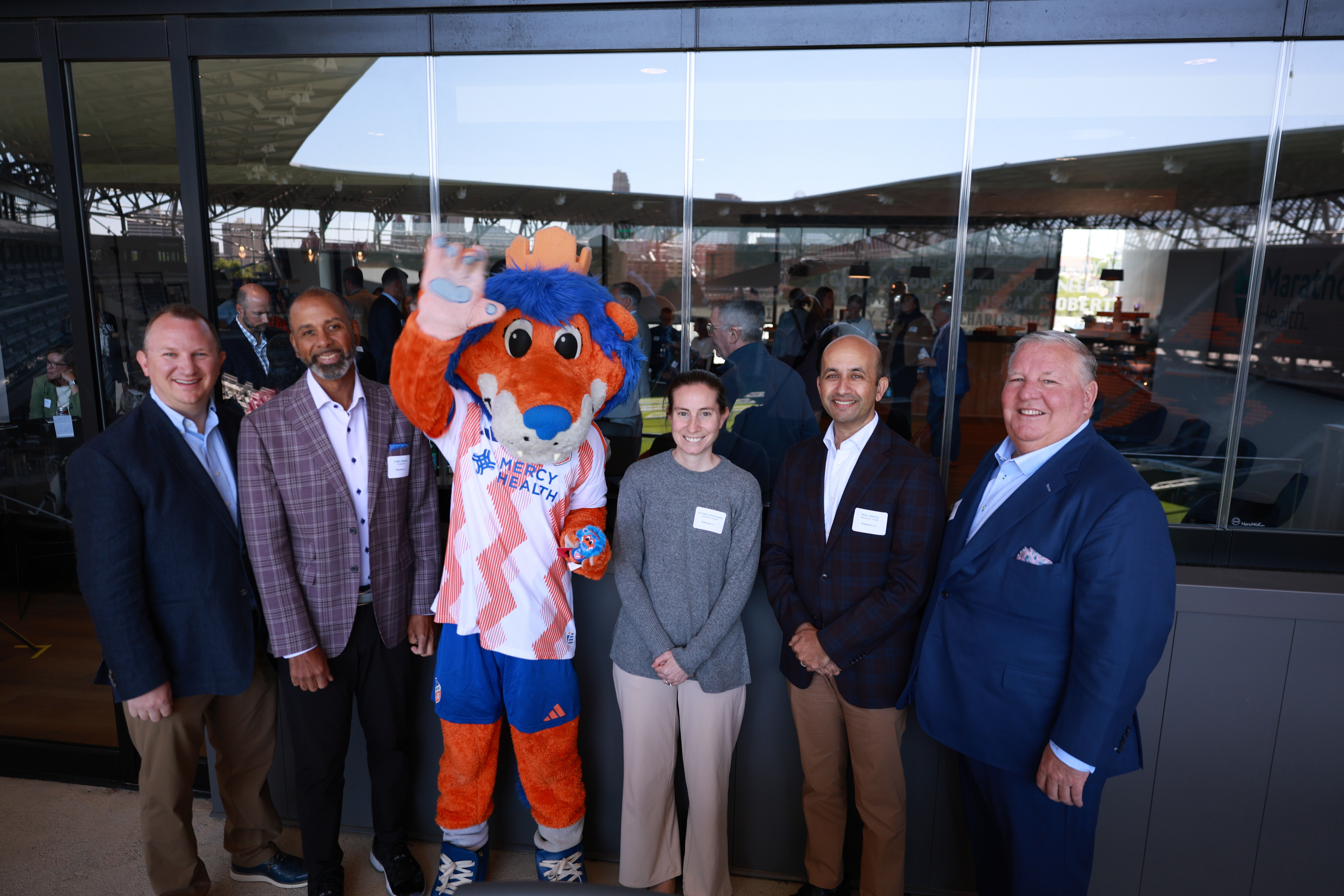 Panelists Matt Mikolajewski, Marc Watkins, Evelyn Hemmingsen, Nirav Vakharia, and Jamie Smith pose with the Mercy Health mascot at TQL Stadium.