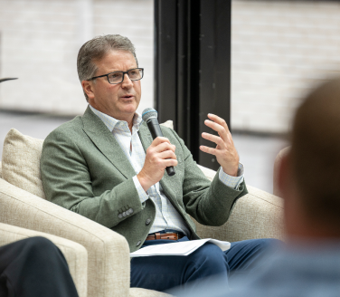 Chris Pricco, chief executive officer of Marathon Health, wears a sage-green blazer and glasses. He gestures while speaking into a microphone at a Marathon Health panel on advanced primary care and behavioral health in Detroit.