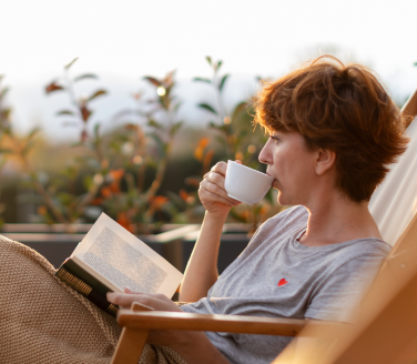 Woman sipping from a cup while reading a book on a terrace.