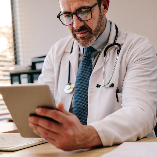 Primary care provider in white scrubs and a stethoscope reviews specialty referral data on a tablet in a bright clinic.