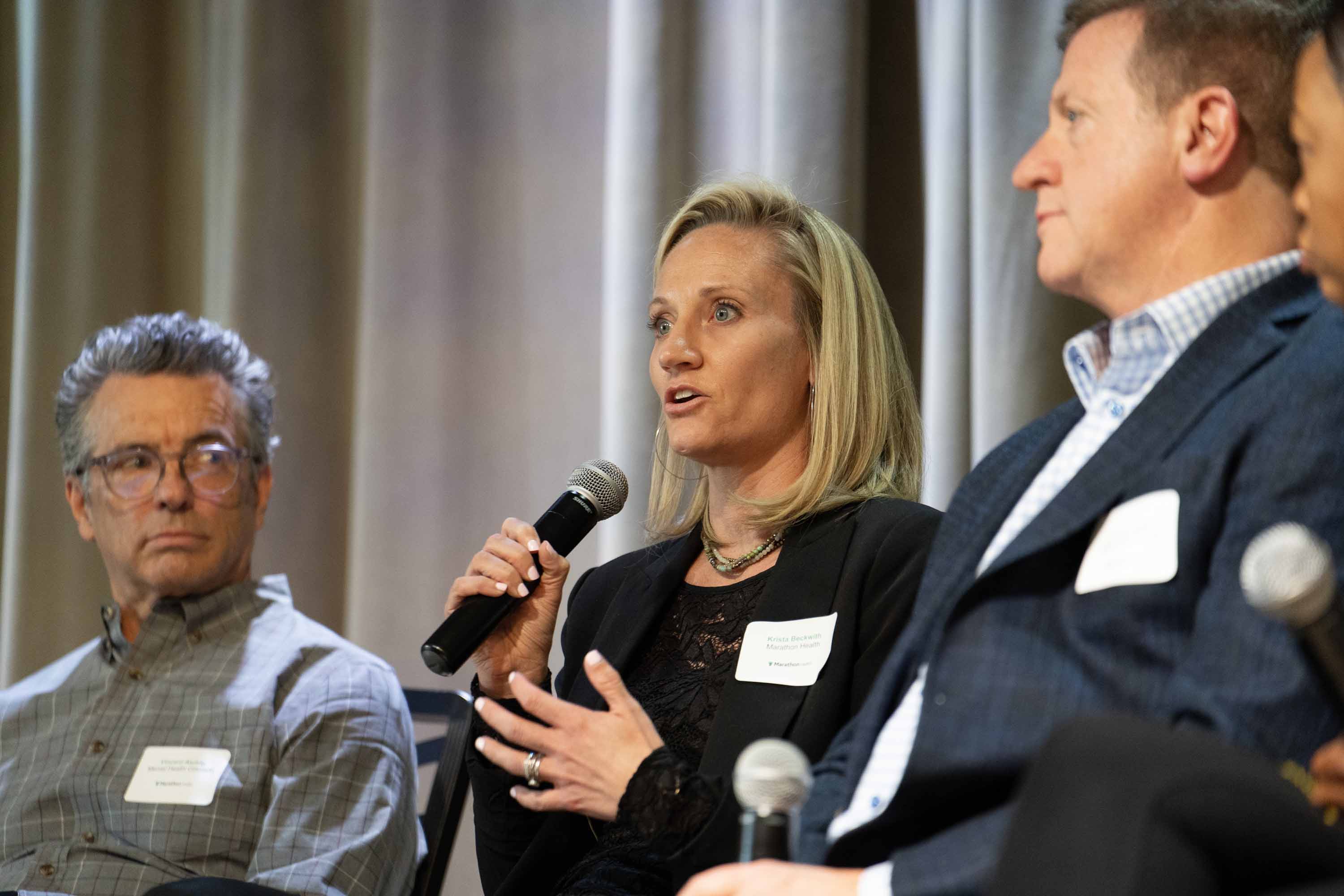 Krista Beckwith speaking into a microphone on stage sitting in between other speakers on stage.