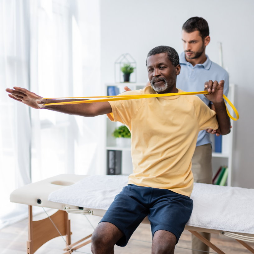 A man does resistance band exercises with arms extended while a physical therapist assists from behind.