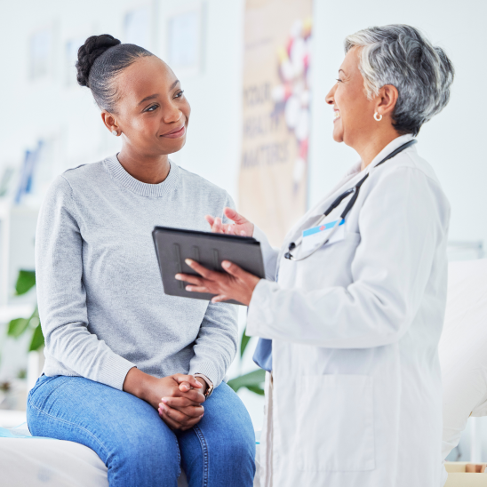 A female doctor with a tablet chats warmly with a young patient sitting on an exam table.