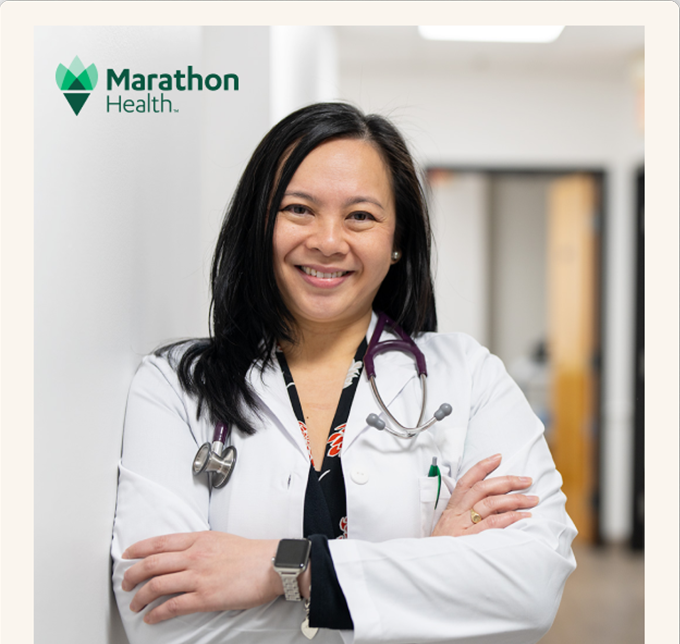 A smiling Marathon Health physician in a white coat stands with arms crossed in a clinic hallway.