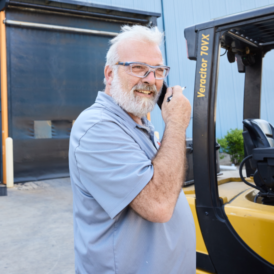 Smiling warehouse worker in safety glasses takes a call by a forklift.